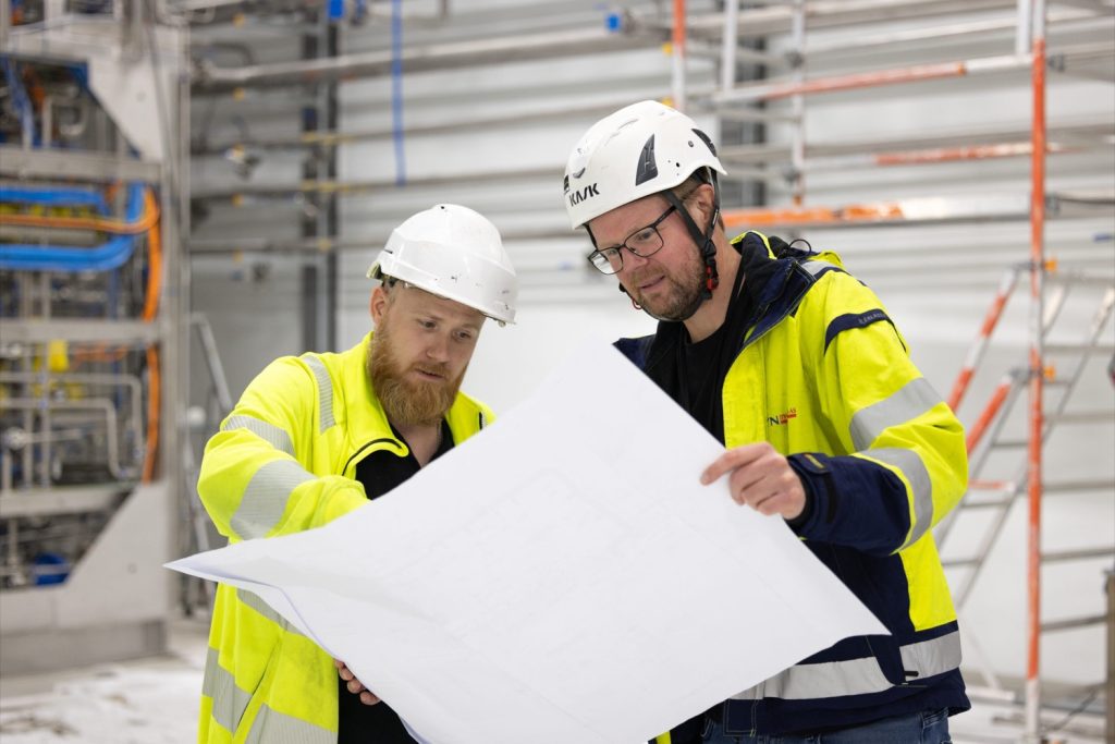 To montører med hjelm som ser på en tegning i et teknisk rom